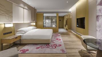 A modern hotel room with a large bed, bedside tables, a desk, a chair, a TV, and a bathroom visible through a glass door.
