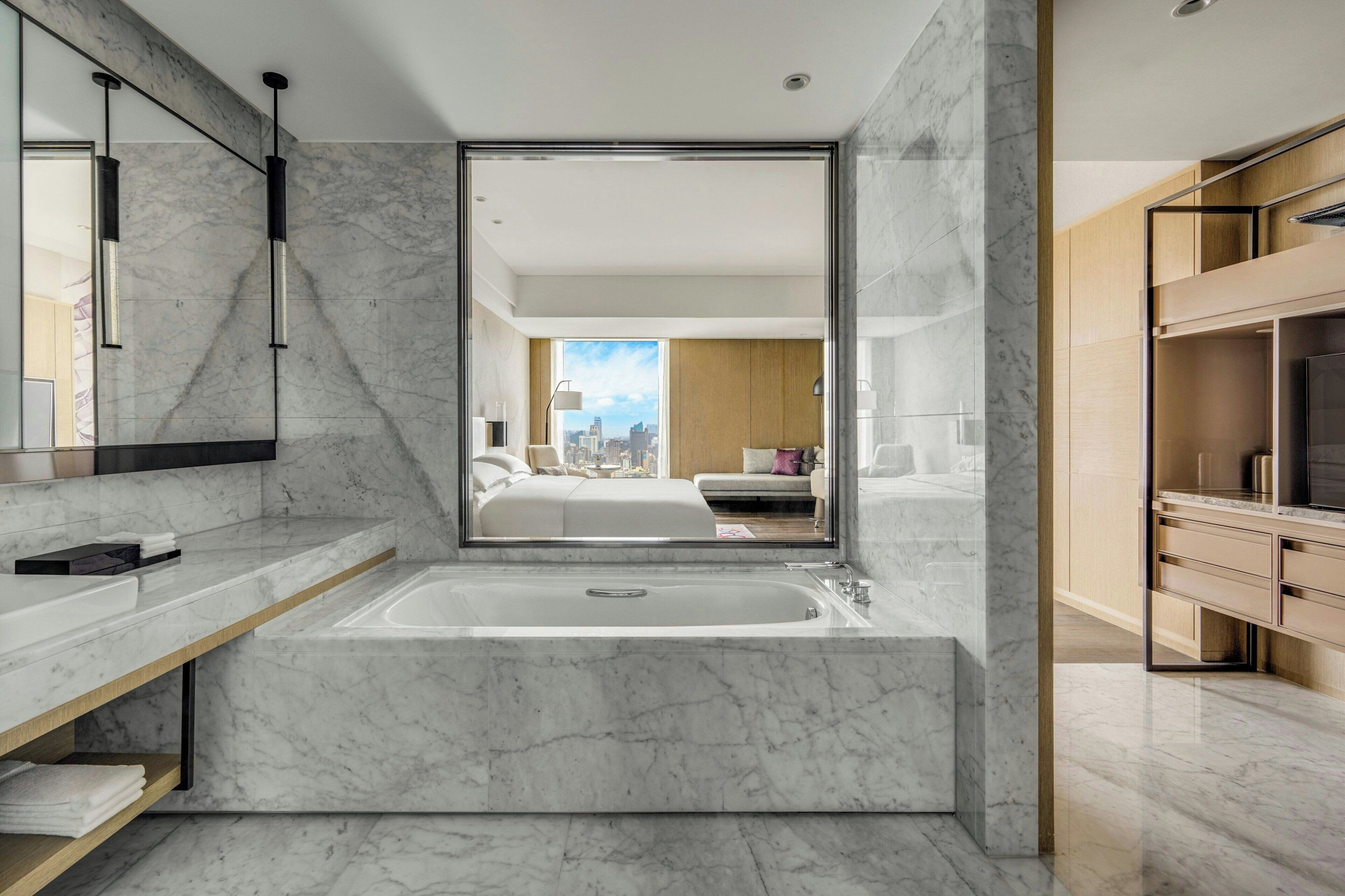 A modern bathroom with a large bathtub, marble walls, and a view of a bedroom.