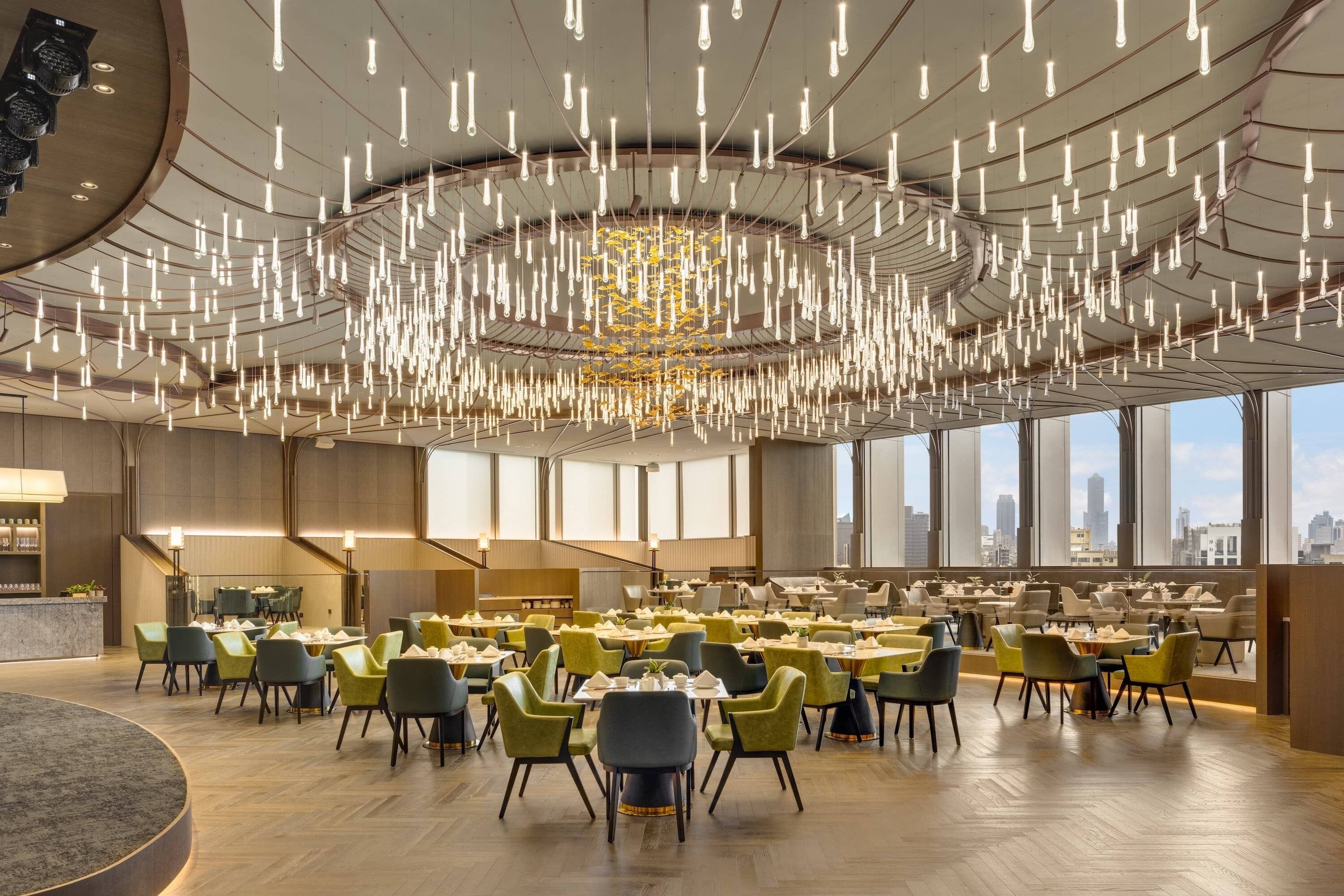 A spacious dining area with multiple tables and chairs, large windows offering city views, and a modern chandelier.