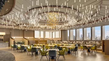 A spacious dining area with multiple tables and chairs, large windows offering city views, and a modern chandelier.