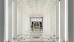 Un largo pasillo simétrico con suelo reflectante y columnas blancas, que conduce a una entrada imponente con una araña.