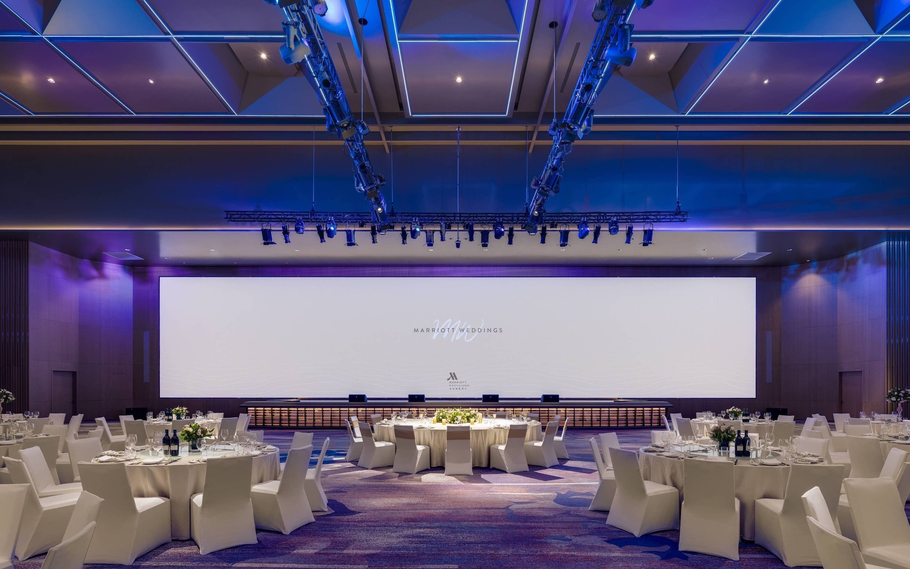 Une grande salle de conférence dotée d’une scène, de rangées de tables rondes recouvertes de nappes blanches et de chaises.