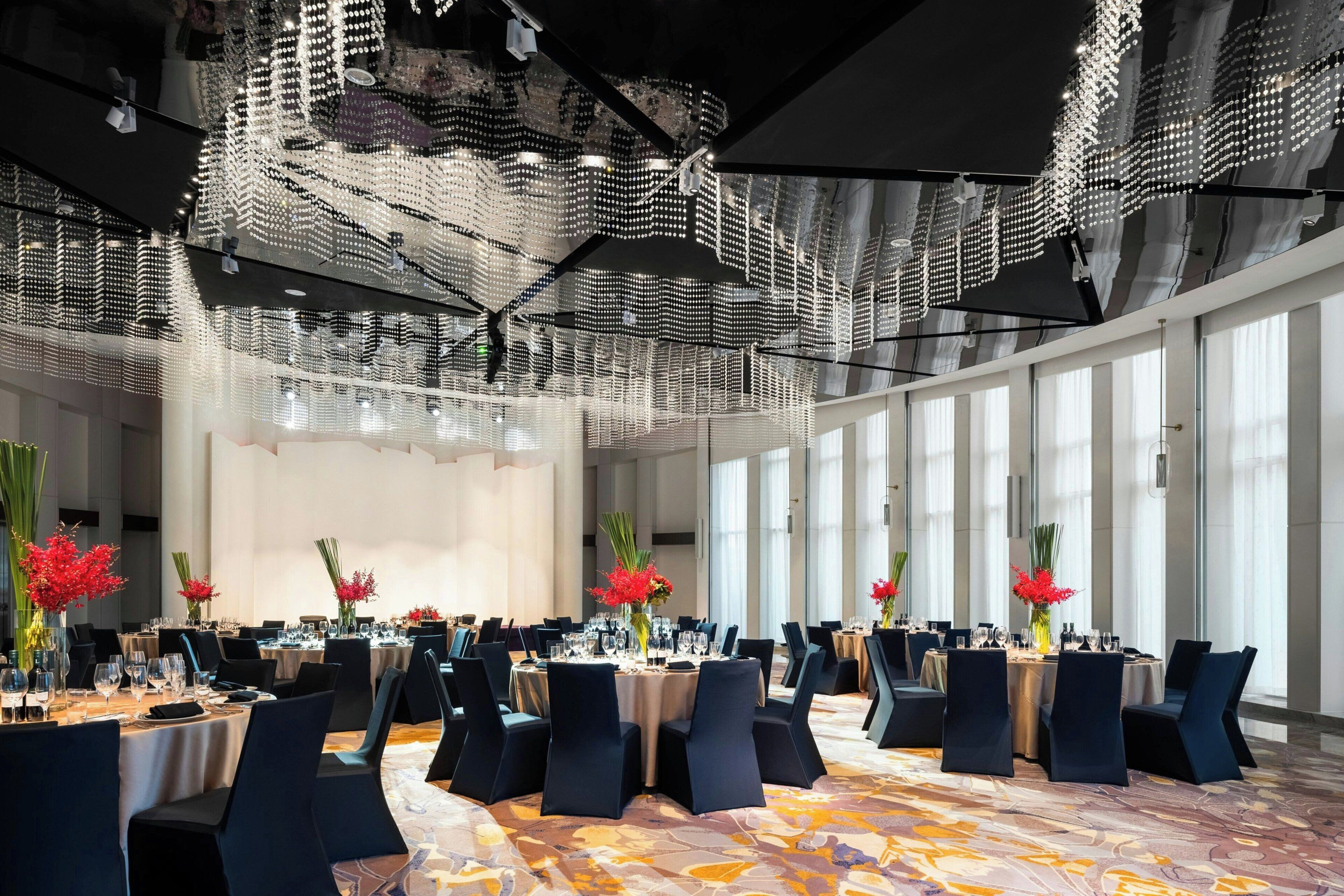 A spacious banquet hall with multiple round tables, dark chairs, and a ceiling with a unique light fixture.