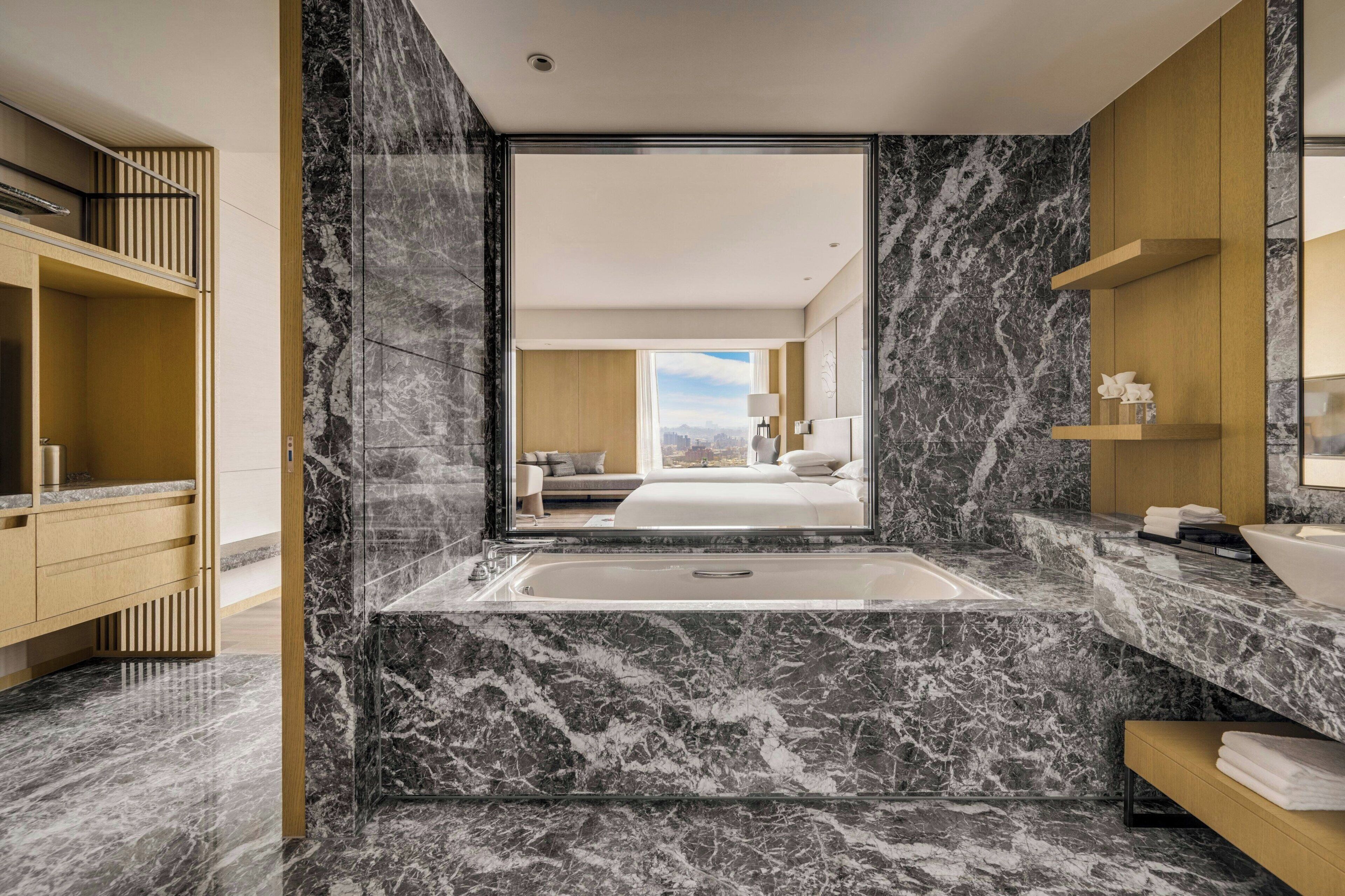A modern bathroom with a marble floor, a large bathtub, and a view of a bedroom with a bed and cityscape.