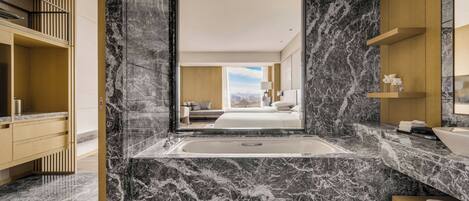 Un baño moderno con suelo de mármol, una bañera grande y vistas a un dormitorio con cama y paisaje urbano.