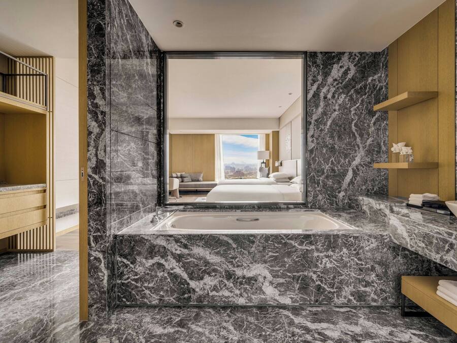 A modern bathroom with a marble floor, a large bathtub, and a view of a bedroom with a bed and cityscape.