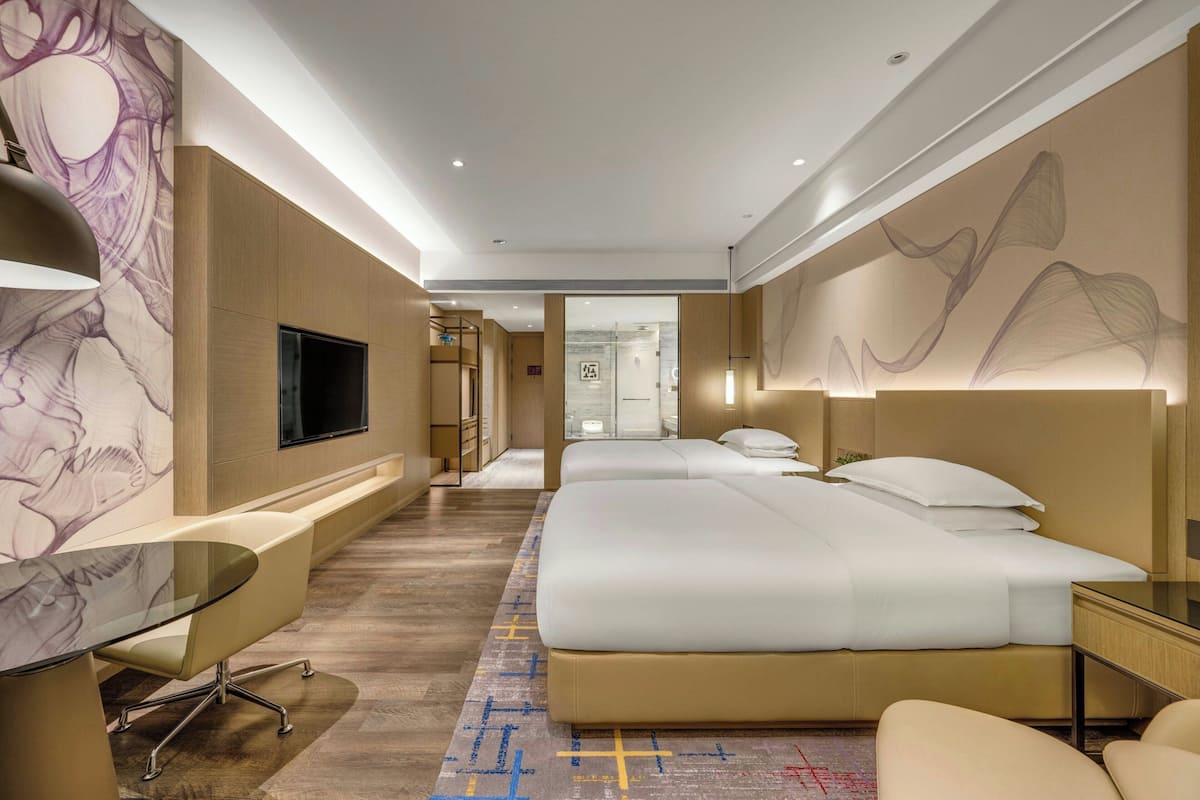 Una habitación de hotel con cama, mesitas de noche, televisor y zona de estar con una mesa de vidrio.