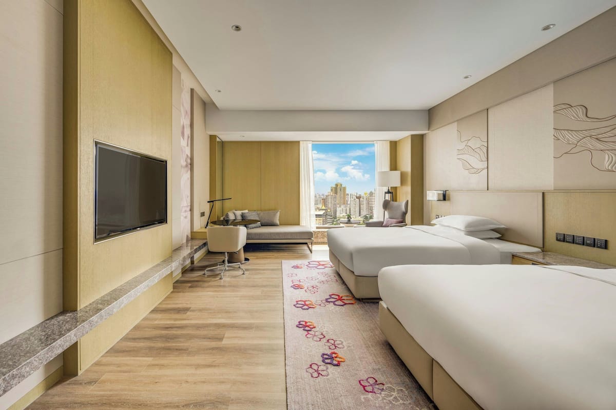 Habitación de hotel con una cama grande, una cama más pequeña, un televisor, un escritorio y vistas a la ciudad.