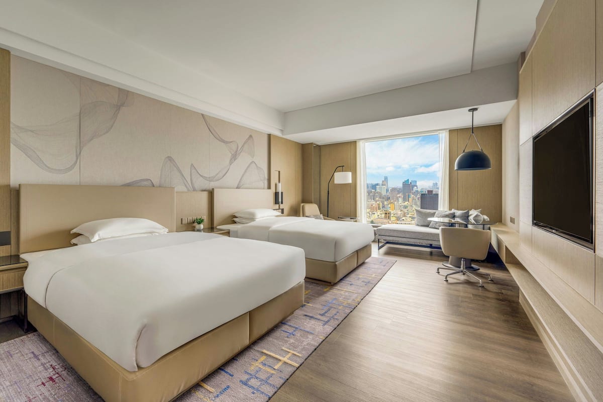 Habitación de hotel con dos camas, un escritorio, una silla, un ventanal grande con vista a la ciudad y un televisor de pared.