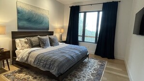 Un dormitorio con cama, ventilador de techo, ventana grande con cortinas y televisor montado en la pared.