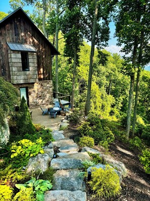 A rustic cabin with a stone exterior, a patio with outdoor furniture, and a stone pathway leading to the cabin. - Luxury Mountaintop 5 min. to Downtown  New Large Modern Mountain design w VIEWS (Asheville)