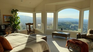 Un dormitorio amplio con una cama grande, una silla, un reposapiés y vistas a la ciudad a través de ventanas amplias.