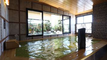 A traditional Japanese onsen with a wooden deck, large windows offering a view of a landscaped garden, and a stone wall on one side.