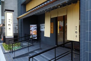Un restaurante con fachada moderna, que destaca por su revestimiento de baldosas y un toldo amarillo. Se observan letreros con caracteres japoneses y un pequeño espacio al aire libre con una planta verde.
