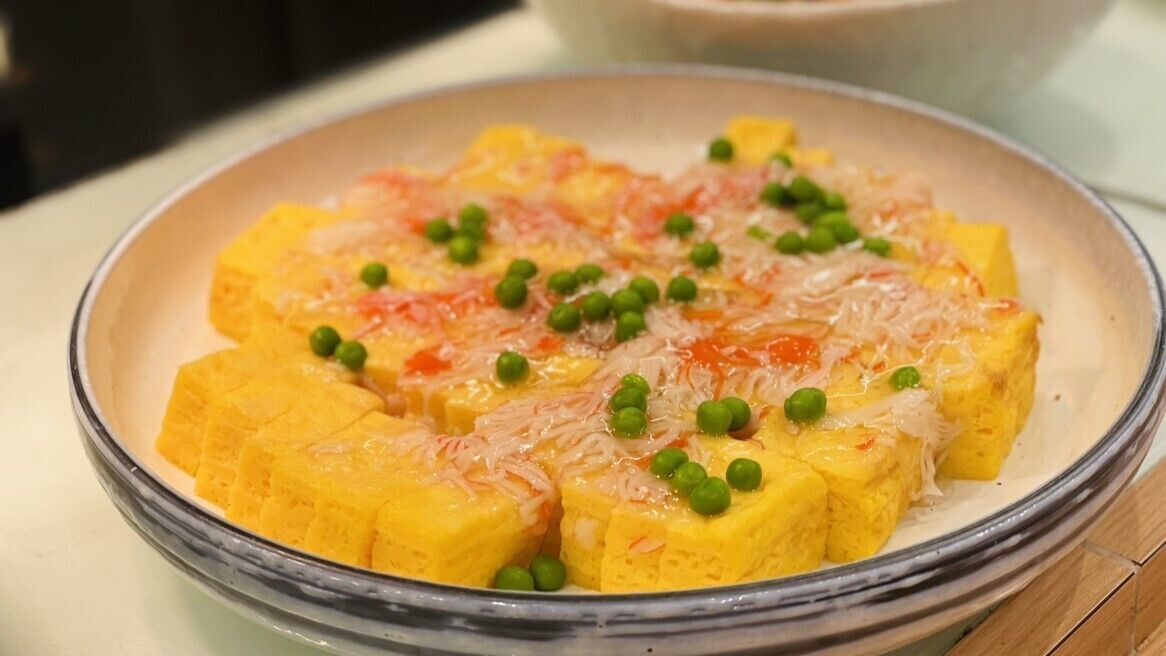 A dish with yellow cubes, green peas, and shredded topping on a plate.