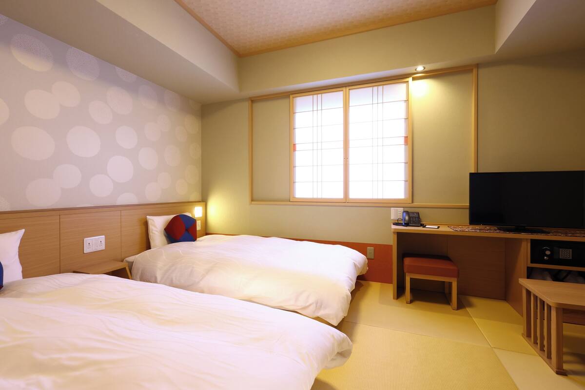 Habitación de hotel con dos camas, un escritorio, televisión y una ventana con persianas.