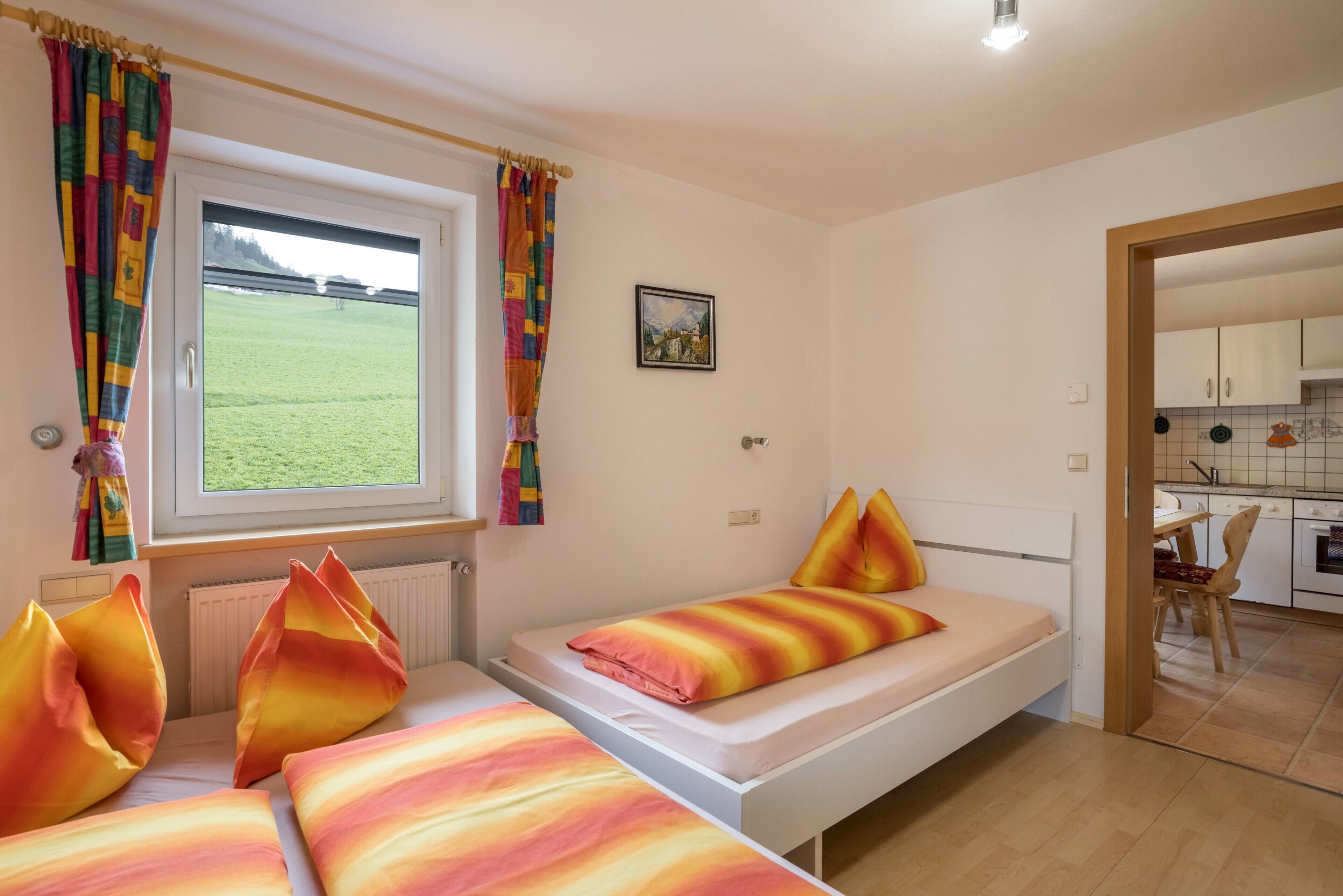 Une chambre avec un lit simple, une fenêtre avec des rideaux, une vue sur un champ verdoyant et une cuisine visible à travers une porte ouverte.