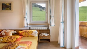 Een slaapkamer met een bed, een raam met gordijnen, een radiator, een nachtkastje met een boek en uitzicht op een groen landschap.