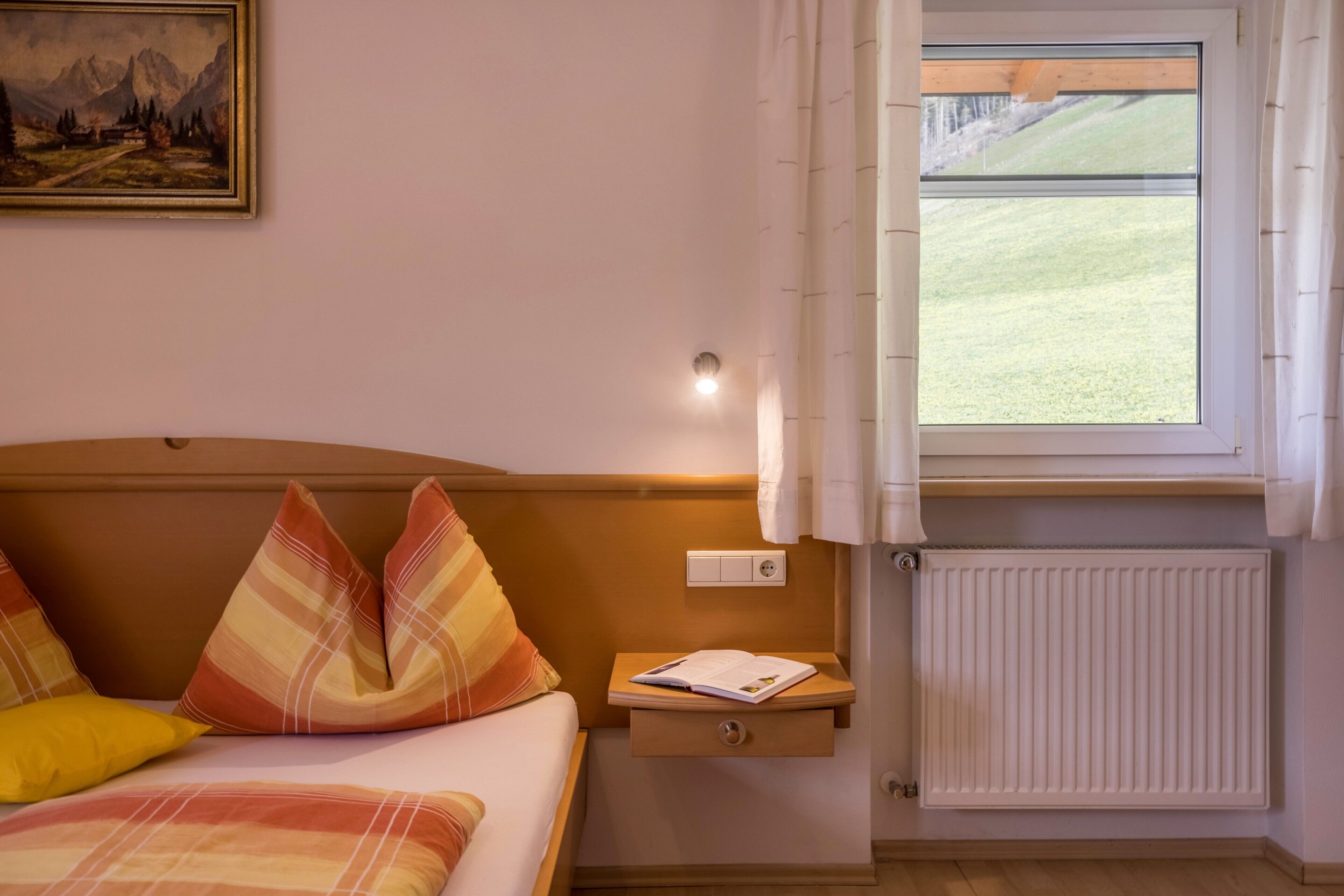 A bedroom with a bed, pillows, a nightstand with a book, a wall-mounted light, a window with curtains, and a radiator.