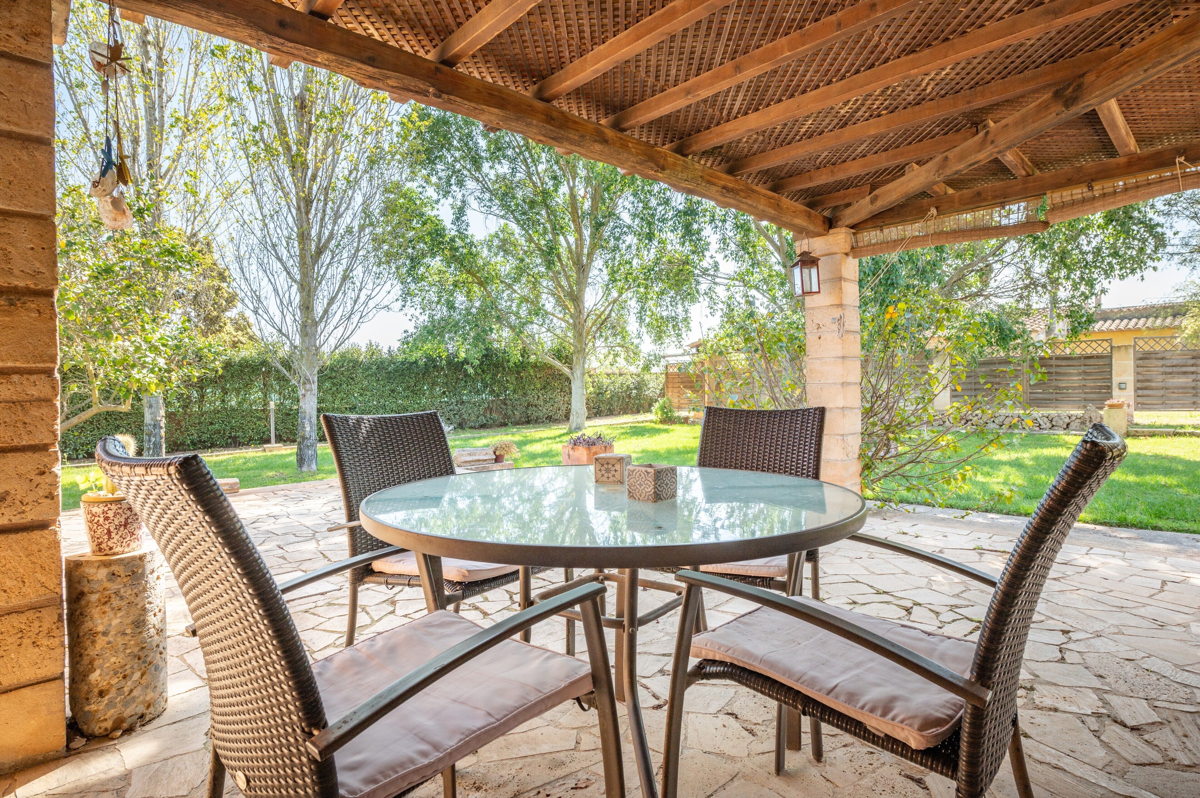 Un patio exterior con una mesa redonda de vidrio y sillas de mimbre, rodeado de muros de piedra y follaje.