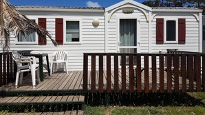 A white mobile home with red shutters, a wooden deck, and a thatched roof structure.