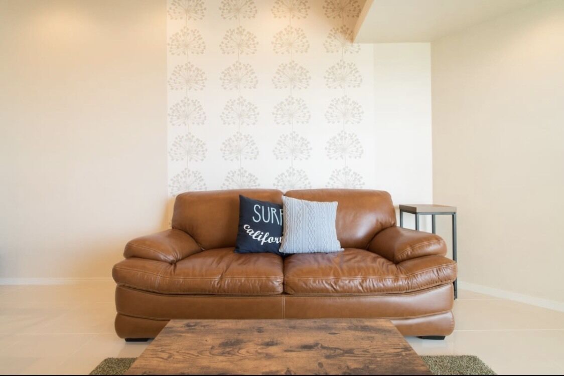 A brown leather sofa with two pillows, one with text, in a room with a patterned wallpaper and a small side table.
