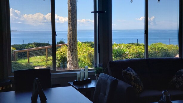 A room with a large window overlooking a coastal landscape, a table with a laptop, and a sofa with cushions.