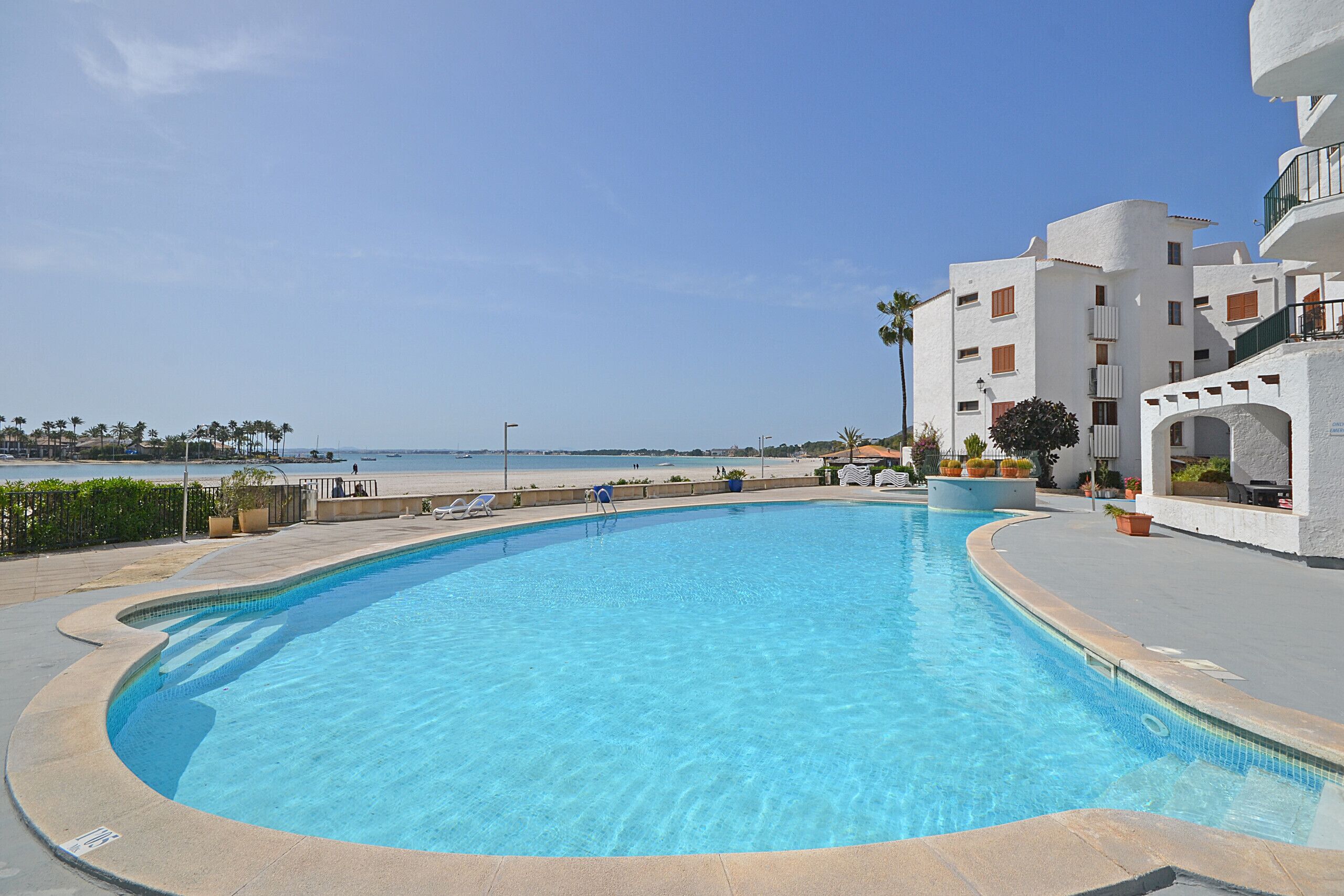 Uma piscina azul límpida com borda curva, rodeada por um muro de pedra baixo. Existem espreguiçadeiras ao longo da piscina, e um edifício com varandas e janelas ao fundo.