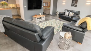 Una sala de estar moderna con un sofá de cuero negro, una mesa de centro y un televisor. Se observa una cocina al fondo, con salpicadero de azulejos y un televisor de pantalla plana sobre la encimera.