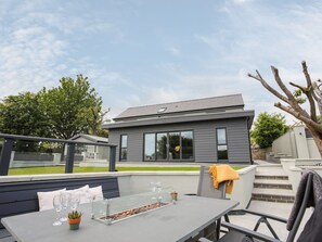 A modern outdoor patio with a glass-top table, seating, and a fire feature. - Maes Llwyd (Pentraeth)