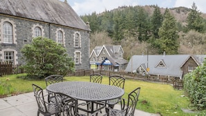 A stone building with a traditional design, a fenced garden, and a table with chairs in the foreground. - Ty Capel (Betws-Y-Coed)