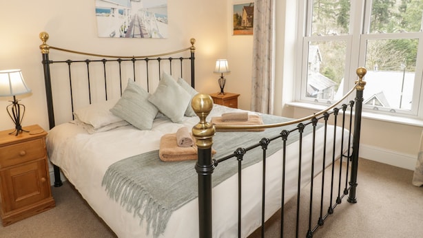 A neatly made bed with a headboard, a bedside table with a lamp, a window with a view of trees, and a painting of a lighthouse on the wall. - Ty Capel (Betws-Y-Coed)
