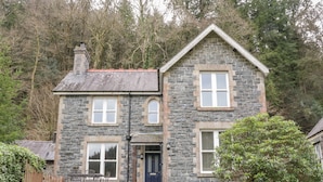 A two-story stone house with a dark blue door, white-framed windows, and a small front porch with chairs. - Ty Capel (Betws-Y-Coed)