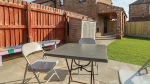 A backyard with a patio table and chairs, a wooden fence, and a brick house. - Rose (Selby)