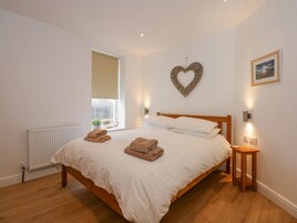 Un dormitorio con cama de madera, ropa de cama blanca, un adorno en forma de corazón en la pared, una mesita de noche pequeña y una ventana con vistas.