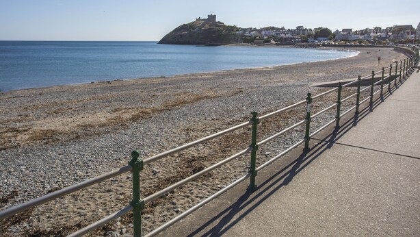 A coastal promenade with a pebble beach, a metal railing, and a distant hill with buildings.