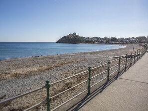 A coastal promenade with a pebble beach, a metal railing, and a distant hill with buildings.