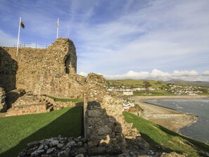 A historic stone structure with a flag, overlooking a coastal town with houses and a beach. - Galwad Y Mor (Criccieth)