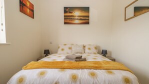 A neatly made bed with a sun pattern bedspread, two bedside lamps, a nightstand, and a painting of a sunset on the wall. - The Croft Bungalow (Wigton)