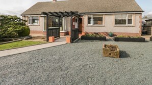A two-story house with a covered porch, a brick chimney, and a gravel driveway. - The Croft Bungalow (Wigton)