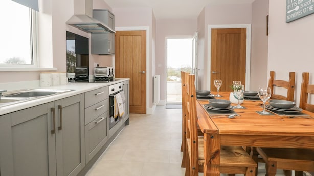 A modern kitchen with grey cabinets, a wooden dining table set for four, and a view to the outside. - The Croft Bungalow (Wigton)
