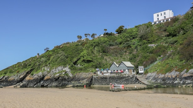 A beach with a building on a hill, a small dock, and a sandy shore. - Eloen (Newquay)