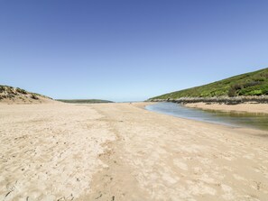 A wide sandy beach with gentle waves, a clear sky, and green hills in the background. - Eloen (Newquay)