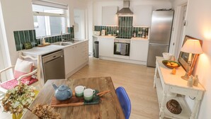 A modern kitchen with white cabinets, a stainless steel refrigerator, and a wooden dining table with a teapot and cups. - Eloen (Newquay)