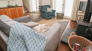 Een woonkamer met een bank, fauteuil en een open haard. Er is een keuken met houten kastjes en een spoelbak.