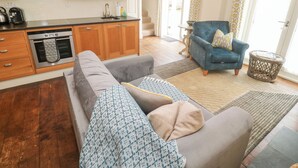 Een moderne keuken met houten kastjes, een inbouwoven en een roestvrijstalen afzuigkap. In de woonruimte staat een grijze bank met decoratieve kussens, en een blauwe fauteuil met kussen. Er is een rieten bijzettafeltje te zien.