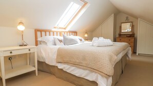 Een slaapkamer met een schuine kap, een dakraam, een bed met een sprei, een nachtkastje met een lamp, een dressoir en een spiegel.