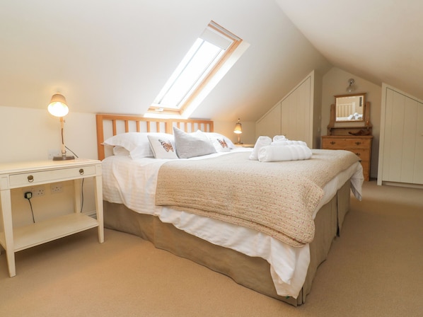 A bedroom with a sloped ceiling, a skylight, a bed with a quilt, a bedside table with a lamp, a dresser, and a mirror.