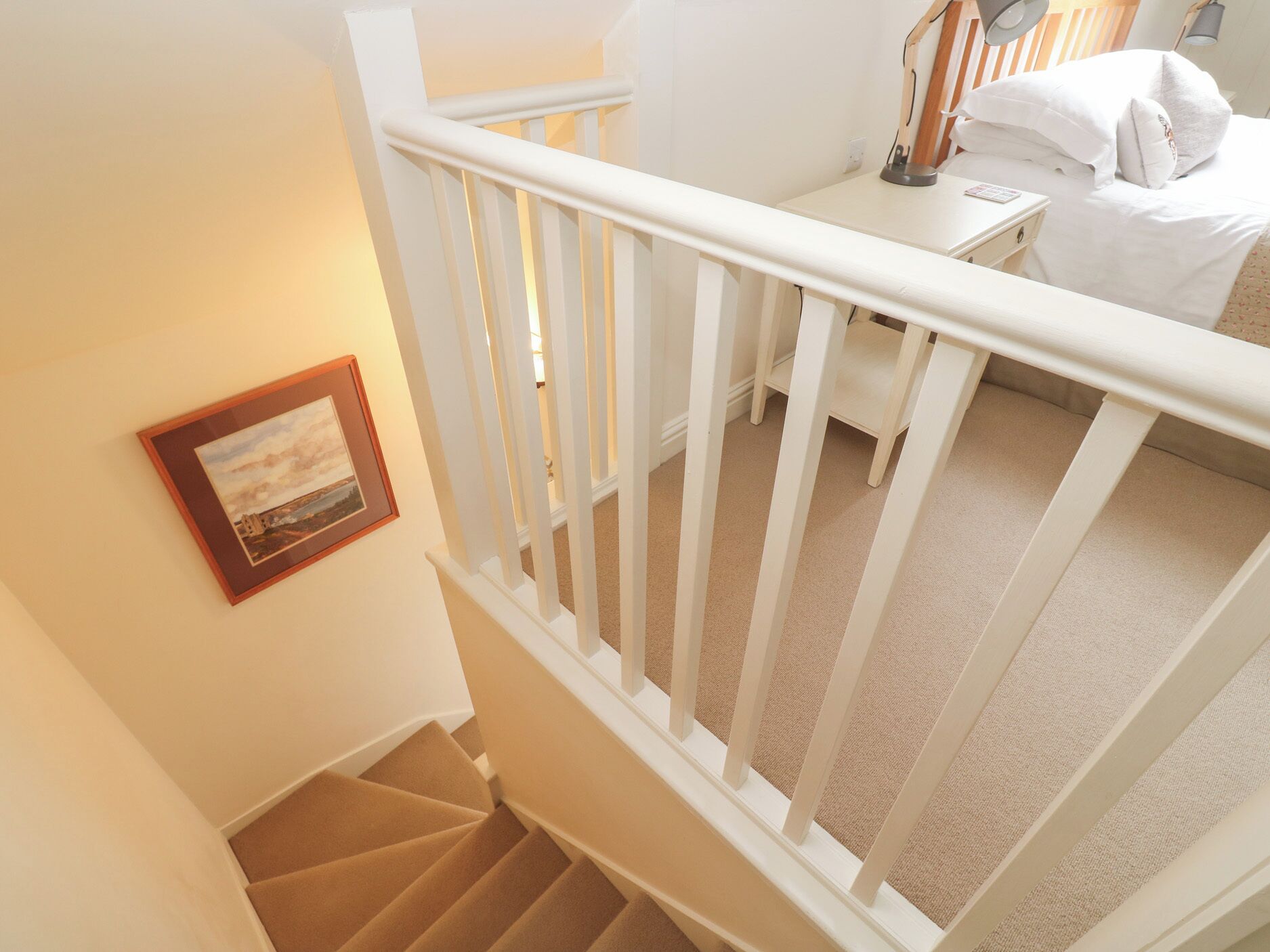 A staircase with white railings leading to a bedroom with a bed, bedside lamp, and a framed picture on the wall.