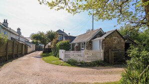 A rural road leading to a stone house with a white picket fence, surrounded by greenery and a gravel driveway. - Toad Hall Cottage (Totnes)